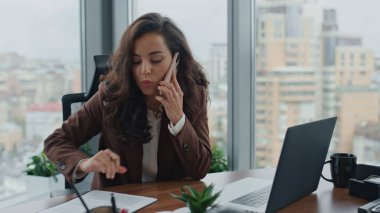 Zeki esmer kadın yönetici, çalışma masasındaki belgeleri kontrol etmek için telefonla konuşuyor. Kendine güveni tam bir iş kadınının ofis masasında telefonla konuşması. Başarılı CEO iş arkadaşını arıyor..