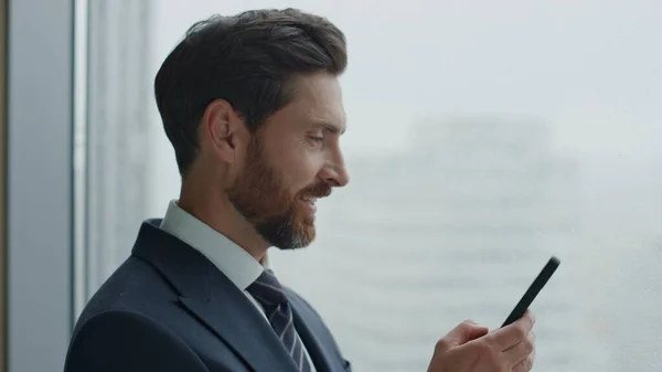 stock image Happy office manager using smartphone sending message standing near skyscraper window close up. Attractive bearded ceo businessman typing email on modern phone. Satisfied man smiling reading news.