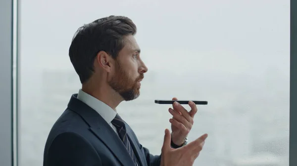 Confident bearded man speaking into speaker phone standing near office window close up. Side view of attractive serious manager have phone conversation using loudspeaker. Smart businessman working.