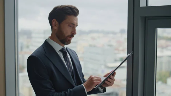 Serious tired businessman checking corporate papers standing at office window close up. Ceo manager reading documents analyzing financial paperwork. Middle-aged bearded man worried about business.