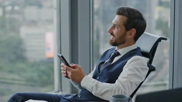 Satisfied entrepreneur watching phone receiving sms sitting at office in elegant suit close up. Confident bearded business man reading good news on smartphone screen. Manager taking break from work.