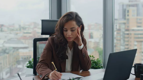 Pensive woman director writing papers sitting at modern table luxury office close up. Thoughtful businesswoman feeling tired overworked working in company. Elegant brunette thinking on business issues