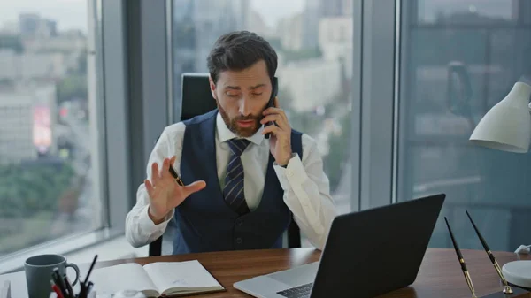 Confident bearded consultant making appointment by phone call noting in notebook at desk. Busy ceo manager using laptop during talking on smartphone. Successful entrepreneur working in modern office.