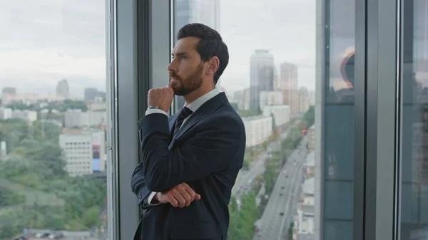 Confident bearded office manager thinking about business ideas standing near window with great cityscape close up. Middle-aged smart businessman creating new company strategy. Employee working alone.