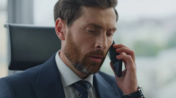 Worried bearded office manager answering call talking by smartphone sitting in modern cabinet close up. Attractive middle-aged businessman have telephone conversation. Ceo entrepreneur feeling tired