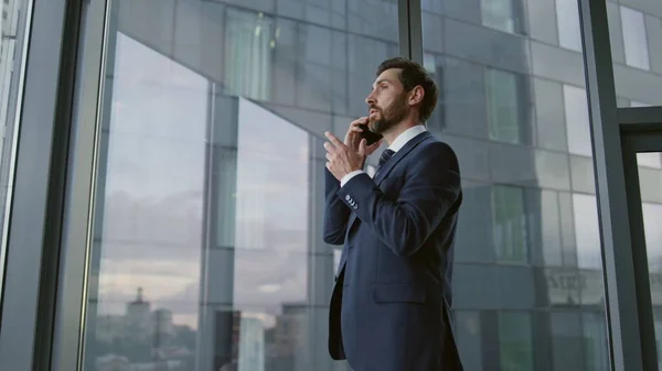 Worried ceo calling phone standing near office window close up. Confident bearded executive manager have serious telephone conversation feeling stress. Busy nervous business man finish smartphone talk