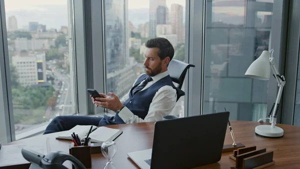 Unsatisfied businessman reading phone message sitting luxury office with panoramic windows. Attractive bearded ceo manager worried about business results. Busy company worker thinking about work.