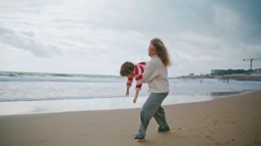 Neşeli anne, sonbahar kumsalında çocuk çeviriyor. Gülümseyen bebek bakıcısı elinde kıvırcık çocukla uçakla oynuyor. Güzel, genç bir anne, oyuncu bir oğul deniz kenarında birlikte eğleniyor. Aile ilişkilerini sevmek