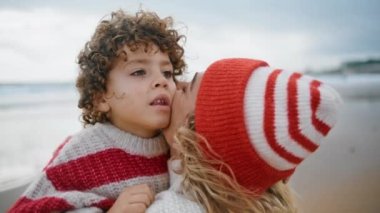 Yakından anne örgü kıyafetli çocuğa sarılıyor. Kumsalda güzel bir aile hafta sonu. Değer veren güzel kadın ebeveyn okyanus kıyısında sevimli kıvırcık çocuğu kucaklıyor. Sevimli çocuk annesiyle vakit geçiriyor.