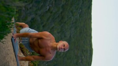 Sabah yeşil dağlarda Asana egzersizi yapan güçlü bir yogi. Odaklanmış adam, akşam dikey çekimde spor minderinde aşağı doğru köpek pozisyonu alıyor. Kaslı yakışıklı adam güzel manzarada üstsüz geriniyor..