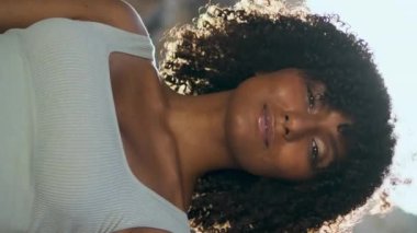 Yaz güneşinin önünde dikey duran Afrikalı Amerikalı güzel bir kız portresi. Yoga eğitiminden sonra Rocky Beach 'te kameraya yakın duran kıvırcık bir kadın. Sakin esmer, güneşli doğada dinleniyor.