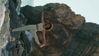 Kayalık deniz kıyısı yaz günü dikey videosunda yoga yapan formda bir kadın. Genç Afro-Amerikan kız tek bacağı üzerinde hatha yoga egzersizleri yapıyor. Sıska kadın eğitim esnekliği