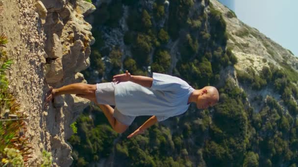 运动员在美丽的海悬崖边集中瑜伽 专注的男人在阳光下做着惊天动地的动作 放松的人运动享受自然和谐在海洋垂直拍摄 禅如天平 — 图库视频影像