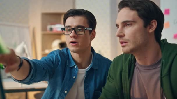 オフィスの近くにコンピュータを指している焦点を当てた同僚 眼鏡の男はパートナーに助言を与える 2人の男が一緒にプロジェクトで働いているPcモニター画面を探しています 締め切りがある深刻なクリエイター — ストック動画
