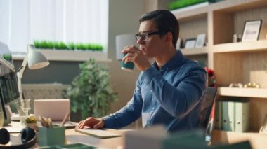 İş yerinde kahvesini yudumlamakla meşguldü. Çağdaş ofiste bilgisayar klavyesi yazan gözlüklü adam kapuçino içiyor. Düşünceli yaratıcı tasarımcı iş yerinde mesaj atma düğmeleri 