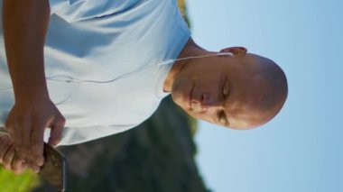 Mutlu adam dağlarda akıllı telefon tutar. Gülümseyen adam podcast 'in tadını çıkarıyor. Açık hava aktivitesi dikey olarak çekiliyor. Tatmin olmuş telefon kullanıcısı tek başına dinlenen kulaklıkları ayarlar. Sosyal medya kavramı.