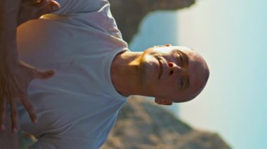 Güçlü adam güzel okyanus kayalıklarında yoga yapıyor. Odaklanmış bir adam, akşam güneşinde Asana pozisyonu üzerinde çalışırken dinleniyor. Atletik kel yogi, deniz manzarasına dikey olarak odaklanıyor..