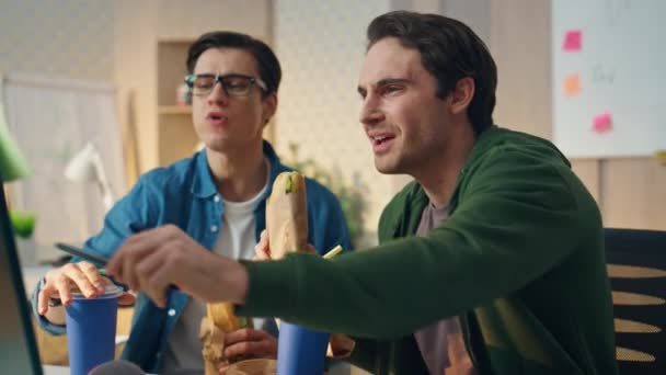 在现代写字楼的特写镜头中 看起来很积极的男人正在监视三明治的吃 笑的家伙指着电脑屏幕 两个饿了的同事正在吃美味的饭菜 队伍共进午餐愉快 — 图库视频影像