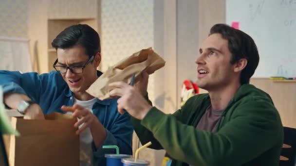 オフィスのクローズアップで同僚にサンドイッチを提案する幸せな男 おいしい食事を食べている2人の男がモニター画面を見ています ランチ休憩を楽しむポジティブな感情を示すフレンドリーなチーム スタッフ グループ 楽しむ — ストック動画
