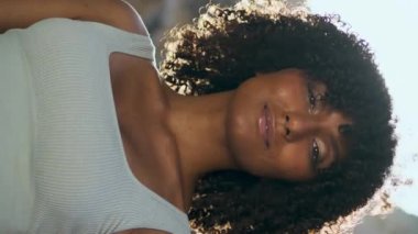 Yaz güneşinin önünde dikey duran Afrikalı Amerikalı güzel bir kız portresi. Yoga eğitiminden sonra Rocky Beach 'te kameraya yakın duran kıvırcık bir kadın. Sakin esmer, güneşli doğada dinleniyor.