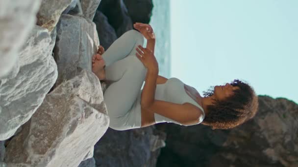 静かな女の子は大きな石の上に座って蓮のポーズをリラックスUrsaビーチ垂直天使 アフリカ系アメリカ人の穏やかな女性の前の岩の海岸で瞑想 若いスポーツウーマンヨガ晴れた日の自然を練習 — ストック動画