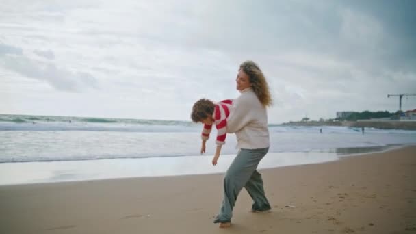 快乐的妈妈在秋天的海滩上纺纱的孩子 笑着照看小孩 手里拿着卷曲的男孩玩飞机 美丽的年轻母亲爱玩的儿子在海滨玩得很开心 关爱家庭关系 — 图库视频影像