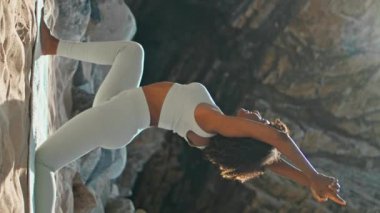 Yüksek kaya dikey videosunun yanındaki kumlu Ursa plajında hilal ay pozu veren Afrikalı Amerikalı kadın. Esnek kız bir dizini kaldırarak savaşçı pozu veriyor. Formda bayan yoga eğitimi alıyor.
