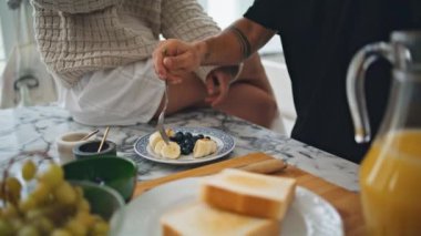 Kahvaltıyı yakın planda yapan çift el. Çatal tutan dövmeli adam tabaktan muz almaya çalışıyor. Bar tezgahında oturan kimliği belirsiz kadın. Evli aşıklar hafta sonu evde yemek yiyorlar.