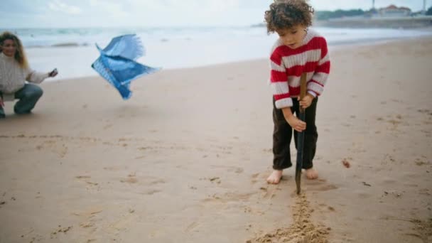 秋の海辺でかわいい子供の砂を描く ビーチで一緒に楽しい家族の遊び 美しい笑顔の母親は息子と週末を過ごすカイトおもちゃを保持 子供の頃の幸せのコンセプト — ストック動画