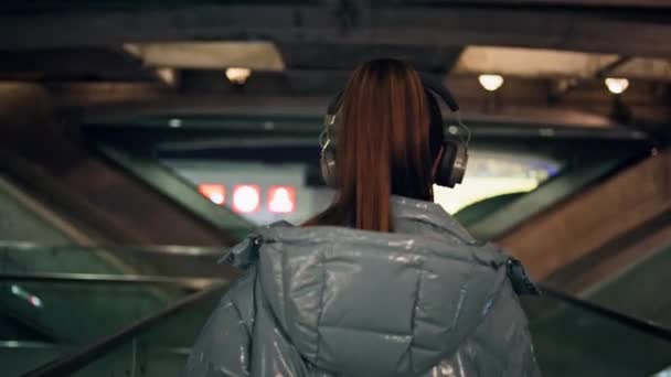 ワイヤレスヘッドフォンで暗い地下鉄を歩いている若い女性のバックビュー カメラに顔を向けるハッピーケアフリーモデルは 笑顔で音楽を聴くことを楽しんでいます ブルネット ロリポップ 楽しい — ストック動画