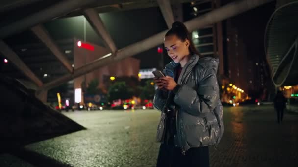 フロントナイトシティに立っているスマートフォンを使用して魅力的な少女ティーンエイジャー 夜の町の散歩でソーシャルネットワークでチャットするケアフリーの若い女性 かわいい学生 笑顔読書メッセージ 屋外で — ストック動画