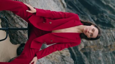 Seksi kırmızı elbisesiyle kayalık sahilde oturan baştan çıkarıcı bir manken. Plaj taşlarında dikey çekimde sandalyede poz veren çekici bir kadın. Güzel, şık bir kız kameraya özgüvenle bakıyor. Güzellik konsepti.