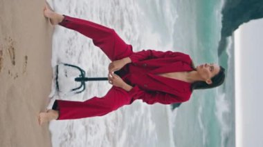 Sahilde oturan şık bayan köpüklü dalgalarla çıplak bacakları yıkadı. Kaya grisi dramatik okyanus manzarasının yanındaki sandalyede poz veren kendine güvenen çekici bir model. Kırmızı şık takım elbiseli şehvetli bir genç kadın..