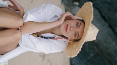 Sahilde oturan mutlu sakin kız geniş hasır şapka takıyor. Kaygısız çekici kadın kameraya gülümsüyor kumlu sahil dikey görüntüsünde dinleniyor. Cazibeli bayan deniz kıyısında tatilin tadını çıkarıyor.