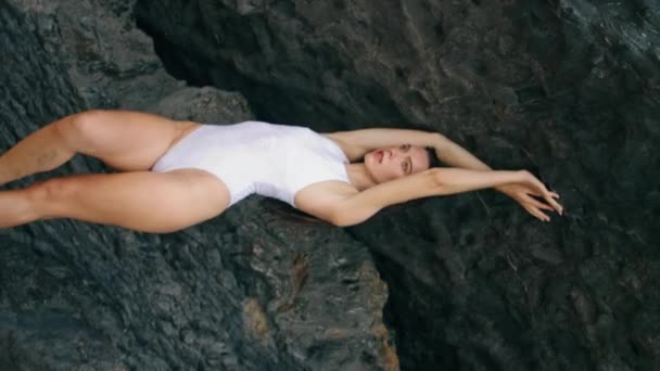 海岸にポーズするセンサモデルは 垂直ビデオをロックする手を上げます 魅力的なセクシーな女性は 石造りの海岸に立っている白い水着を着ています 豪華な深刻な少女はリラックスして夏休みを楽しむ — ストック動画