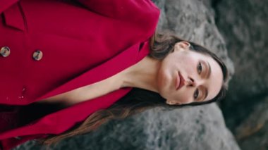 Dağlarda şık kırmızı elbiseli kendine güvenen bir kız. Taş kaya üzerinde poz veren çekici stil sahibi kadın dikey açıdan bakıyor. Ciddi düşünceli bayan kasvetli doğada rahatlar. Moda konsepti