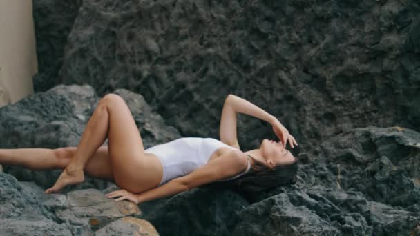 石の岩でリーク海岸にポーズするセクシーな女性 白い水着の魅力的なセンセーショナルモデルは 海岸の垂直方向のビューでカメラを見ています 崖の近くのビーチ砂の上に立っている熱い豪華な少女 — ストック動画