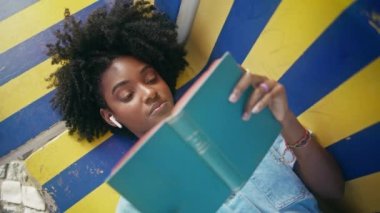 Kapalı kapılar ardında kitap okuyan genç açık havada renkli bir bankta uzanıyor. Kıvırcık Afrikalı kulaklıklı Amerikalı kız edebiyat hobisinin tadını çıkarıyor. Güzel genç bir kadın yaz tatilinde en sevdiği romanla rahatlıyor.