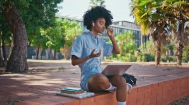 Sunny caddesinde oturan öğrenci bilgisayarla konuşuyor. İnternetteki konferansta konuşan kablosuz kulaklıklı güzel Afro-Amerikalı kız. Kıvırcık moda hipster defter başında sohbet ediyor.