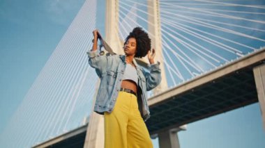 Yaz günü şehir köprüsünün önünde poz veren kaykaylı güzel bir kız. Mavi gökyüzü manzarasında duran havalı Afro-Amerikan genci. Kıvırcık ergen hippi mesafeye bakıyor