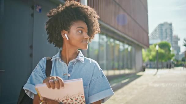大学生去上课 环视阳光灿烂的街道近旁 时尚的非洲裔美国女孩拿着书本走向大学校园 卷曲的女青少年戴着耳机享受夏天的散步 — 图库视频影像