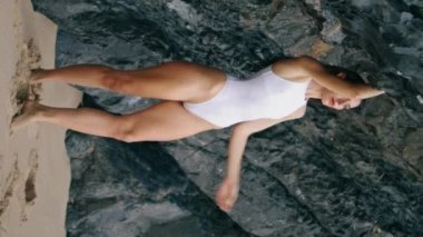 Kum kıyısında ayakta duran seksi kadın dikey kaya görüntüsü için el kaldırıyor. Taş uçurumun kenarındaki sahilde beyaz, zarif mayo giymiş çekici, şehvetli bir model. Muhteşem uzun saçlı kız, dışarıda rahatla.