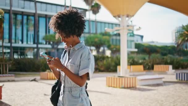 微笑的女人在阳光灿烂的日子里用手机发短信 快乐的非洲裔美国女孩在夏天散步时给智能手机屏幕发短信 可爱的卷曲女士在户外享受远程交流 — 图库视频影像