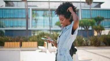 Akıllı telefondan mesajlaşan neşeli genç kız güneşli şehirde yürüyor. Mutlu Afro-Amerikan gençliği cep telefonundan SMS okurken gülümsüyor. Neşeli kıvırcık hippi telefonda sosyal medyayı izlerken gülüyor..