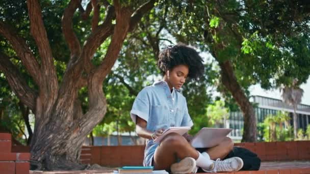 ノートパソコンでオンラインコースを受講する少女は 木の下に座っているコピーブックでノートを作ります ヘッドフォンでウェビナーを聞いているかわいいアフリカ系アメリカ人の学生は コンペディウムを書いています 屋外で勉強する女性ティーンエイジャー — ストック動画