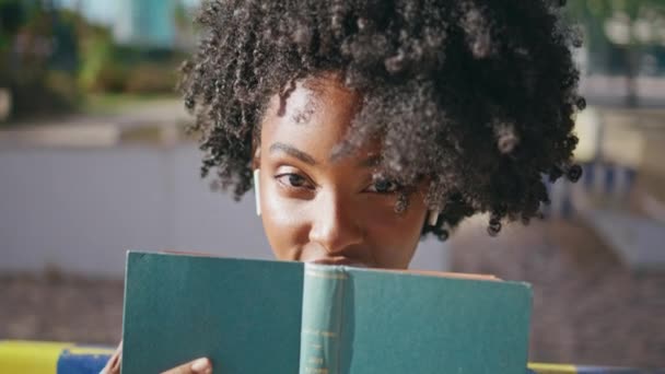 在阳光灿烂的大街上 非洲裔美国学生在漂亮的脸庞前摆姿势看书的画像 迷人的黑发女孩笑着看着相机 可爱的卷曲少年带着最喜欢的小说在户外放松 — 图库视频影像