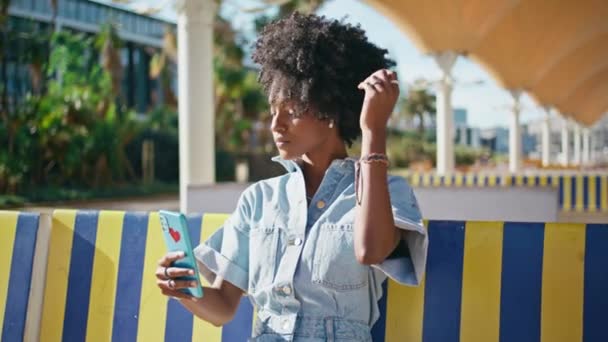 トレンドティーンエイジャーは 屋外のカラフルなベンチに座ってセルフィーを作ります スマートフォンカメラで顔写真を撮る美しいアフリカ系アメリカ人の少女 カーリースタイリッシュなティーン笑顔携帯電話晴れた日 — ストック動画
