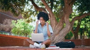 Kulaklık takan genç kız dizüstü bilgisayarlı yeşil ağacın altında oturuyor. Güzel Afro-Amerikan genç doğa üzerine online çalışma için kulaklık takıyor. Akıllı kız öğrenci, bilgisayar dersi alıyor..