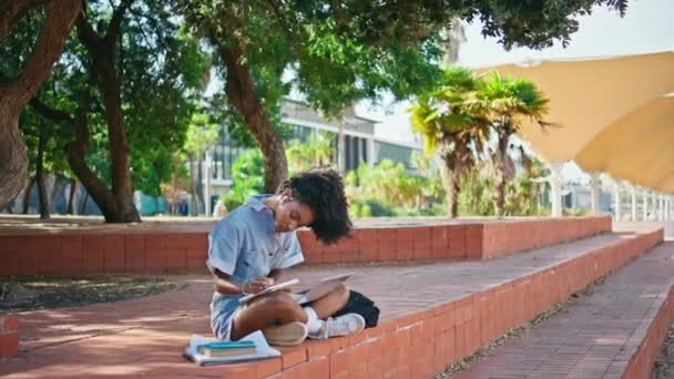 リモートで勉強する大学生は 日当たりの良い通りのコピーブックで宿題を書いています アフリカ系アメリカ人の女の子がノートPcで屋外に座ってノートを作る トレンドの若いヒップスターオンライン学習 — ストック動画