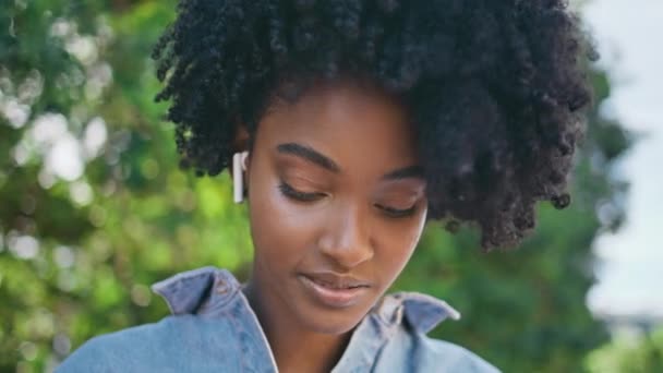 非洲女孩带着无线耳机在网上聊天的肖像 美丽的年轻女子戴着耳机在谈论阳光灿烂的自然 卷曲的黑发学生喜欢远程通信特写 — 图库视频影像